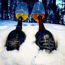 Two Laurent Perrier Grand Siecle champagne bottles partially buried in snow with filled wine glasses on top against a forest backdrop.